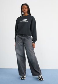 Zwarte sweatshirt met wit "new balance" logo, gecombineerd met losse grijze jeans met lichte slijtage en zwart-witte schoenen.