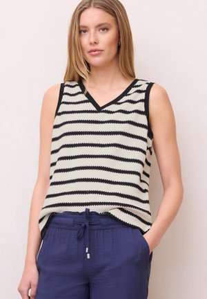 Frau trägt ärmelloses, schwarz-weiß horizontal gestreiftes Oberteil mit V-Ausschnitt und marineblaue Hose mit Kordelzug, steht vor einfarbigem Hintergrund.