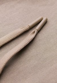 Deux extrémités de lacets tressés beige avec embouts en plastique reposant sur un fond en tissu texturé.