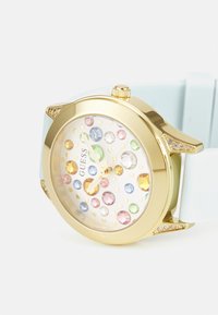 Reloj con caja en tono dorado, esfera blanca con gemas multicolores y una correa de silicona azul clara. Incluye acentos dorados y detalles de cristal.