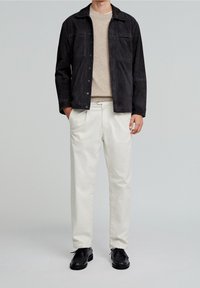 Zwart suède jack over een beige gebreide trui; roomkleurige geplooide broek; zwarte leren schoenen. Geen zichtbare patronen of accenten. Neutrale achtergrond.