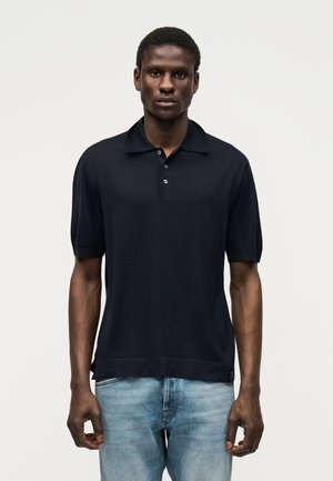 Mężczyzna stojący twarzą do przodu, ubrany w czarną koszulkę polo z krótkim rękawem i jasnoniebieskie dżinsy na jednolitym tle.