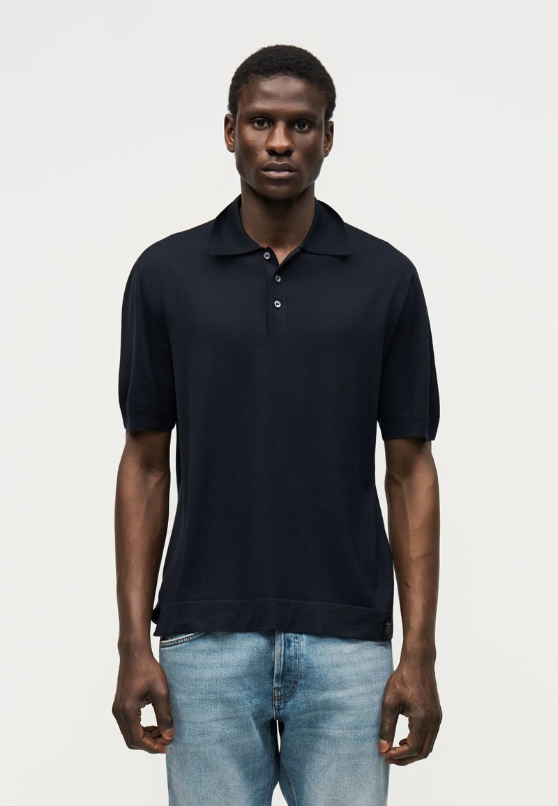 Άνδρας που στέκεται με το πρόσωπο προς τα εμπρός, φοράει μαύρο πόλο με κοντά μανίκια και ανοιχτόχρωμα μπλε τζιν μπροστά σε απλό φόντο.