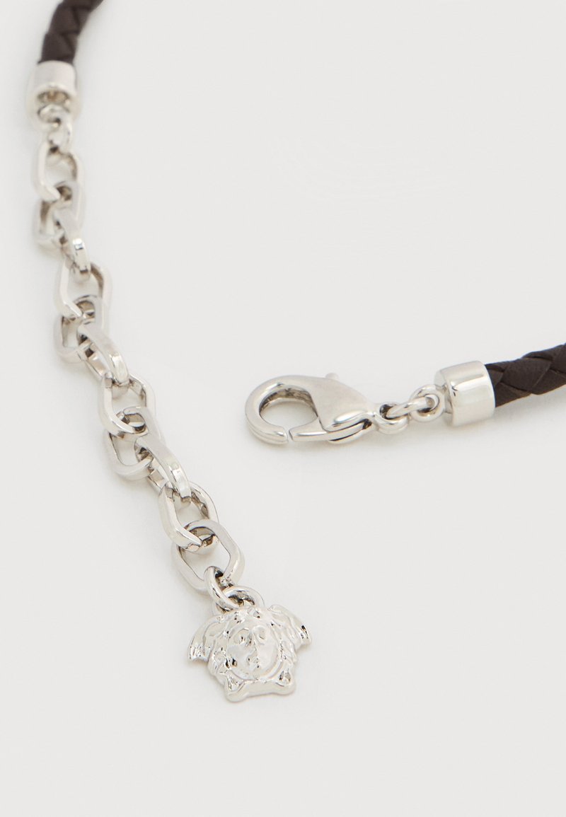 Brauner geflochtener Riemen mit silbernen Beschlägen, versehen mit abgerundeten Gliedern einer Kette und einem Anhänger in Form eines Gesichts. Mit Karabinerverschluss.