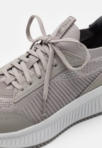 Grå sneaker med ett texturerat nätöverdrag, plana snören och en kontrasterande vit och grå gummisula. Har en "BOSS"-logga på sidan.