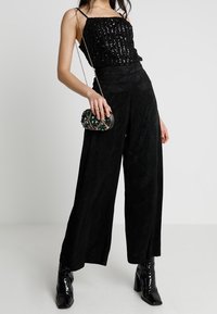 Svart paljett-topp med justerbara axelband, tillsammans med vida svarta byxor med ett texturerat mönster. Tillbehör: liten utsmyckad handväska.