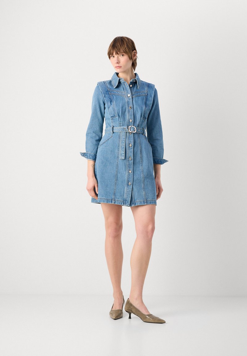 Femme debout portant une robe chemise en jean ceinturée à manches longues et des chaussures pointues à talons bas sur un fond blanc uni.
