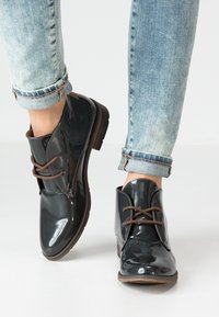 Bottines en cuir verni noir avec des lacets marron, bouts ronds et un petit talon. Portées avec un jean bleu clair retroussé.