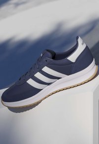 Zapatilla azul marino con rayas blancas y suela de goma, colocada sobre una superficie blanca inclinada bajo luz natural.