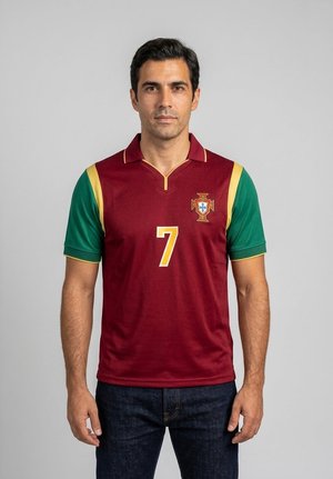 Mann trägt ein dunkelrotes und grünes Portugal-Fußballtrikot mit der Nummer sieben, steht vor einem einfarbigen hellgrauen Hintergrund.