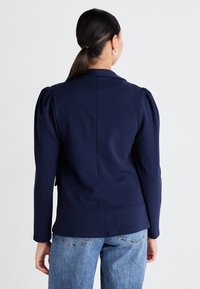 Navy modrý blazer se strukutrovanými rameny a přiléhavým střihem, se zadním švem a dlouhými rukávy, kombinovaný s světle modrými džíny.