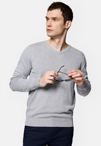 Szary sweter dziany z dekoltem w serek, z prążkowanymi mankietami i dolnym brzegiem, noszony przez mężczyznę trzymającego okulary przeciwsłoneczne, z małym logo na klatce piersiowej.