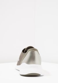 La chaussure de sport Lacoste présente une tige en mesh respirant de couleur vert olive, un talon métallique texturé et une semelle en caoutchouc blanche.