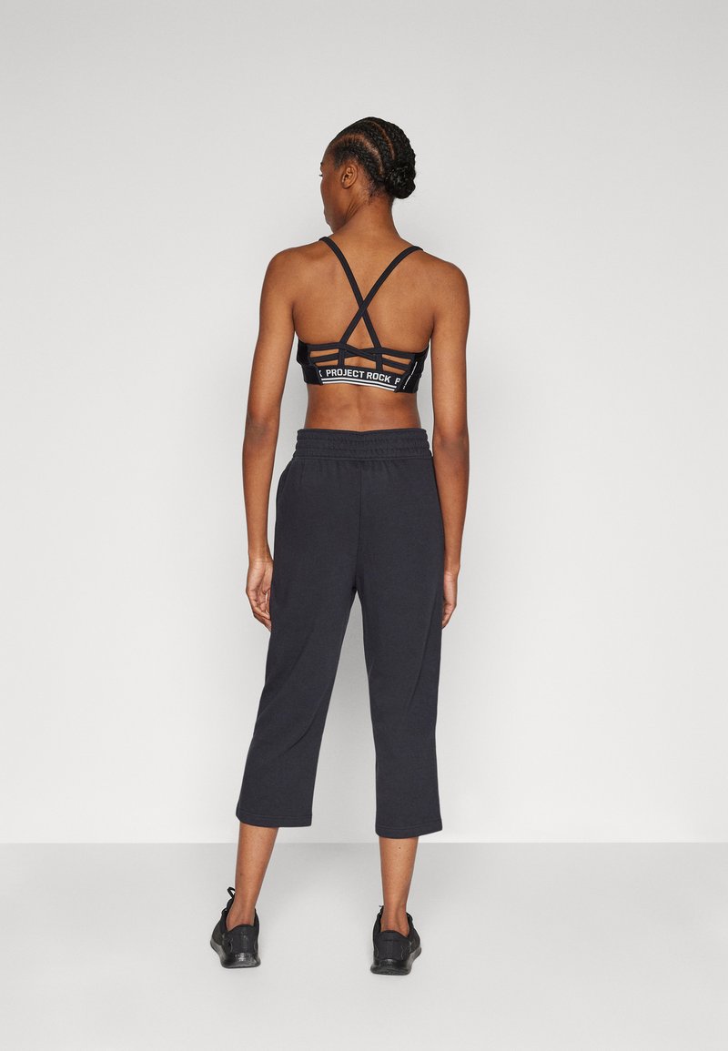 Černá sportovní podprsenka s křížovým designem a logem, spárovaná s černými zkrácenými tepláky a sportovními botami, viděná zezadu.