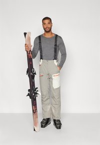 El conjunto de ropa de esquí incluye una camiseta de manga larga gris, pantalones de esquí beige con bolsillos, tirantes negros y botas de esquí negras, sosteniendo un par de esquís.