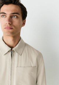 Chaqueta beige texturada con cremallera y cuello tipo camisa, y bolsillo en el pecho. Presenta una cremallera suave y un ajuste estructurado.