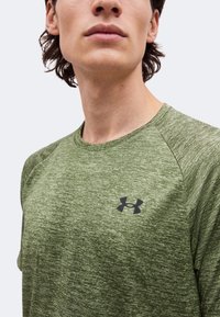 Man wearing a green textured short-sleeve shirt with a black Under Armour logo on the chest, shown from shoulders to chin.