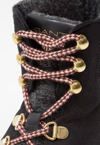 Schwarzer Wildlederstiefel mit grauem Filzkragen, ausgestattet mit kastanienbraunem und cremefarbenem geflochtenem Schnürsenkel, goldenen Beschlägen und Lederakzenten. Nahaufnahme der Ösen.