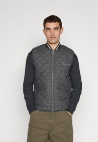 Quilted gray vest with a front zipper, two pockets, contrasting ribbed collar, and embroidered logo on the left chest. Worn over a black shirt.