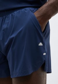 Άτομο με σκουρόχρωμο δέρμα που φοράει αθλητικό σορτς ναυτικού μπλε με ελαστική μέση, το χέρι ακουμπά στην πλαϊνή τσέπη, μικρό λευκό λογότυπο ορατό.