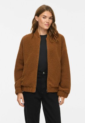 Frau trägt eine braune Fleecejacke mit Reißverschluss über einem schwarzen Top und schwarzen Hosen und steht vor einem schlichten weißen Hintergrund.