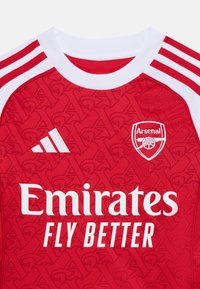 Rød sportsdrakt med hvit krage og ermer, med teksturert mønster, Adidas-logo, Arsenal-emblem og teksten "Emirates Fly Better".