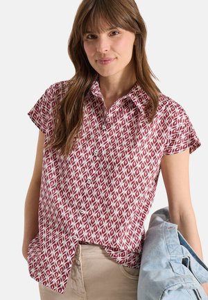 Kobieta w koszuli z krótkim rękawem, zapinanej na guziki, z czerwono-białym geometrycznym wzorem, trzyma jasnoniebieską kurtkę na jednym ramieniu.