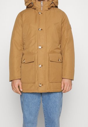 Mand iført en beige hættetrøje vinterjakke med knapper og lommer med klap foran, kombineret med blå jeans foran en ensfarvet baggrund.