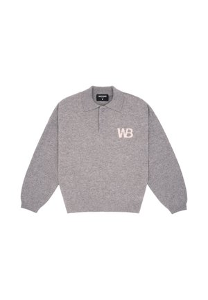 Personne aux cheveux bouclés portant des lunettes de soleil, un t-shirt gris à manches longues avec le logo « WB », un pantalon cargo noir, une chemise à carreaux bleue attachée autour de la taille, et des chaussures noires.