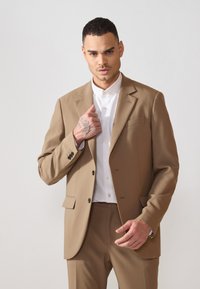 Traje color beige con botones negros y solapa, combinado con una camisa blanca. Textura similar a la seda, ajuste entallado, con líneas elegantes y diseño minimalista.