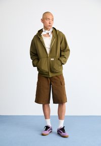 Camisola com fecho éclair em verde-azeitona, top branco, calções de carga castanhos, sapatos cinzentos e roxos, e um lenço branco com padrão ao redor do pescoço.