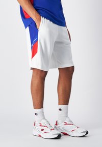 Weiße Sportshorts mit blauen und roten Panelakzenten, kombiniert mit einem blauen Shirt, weißen Sneakers mit roten Details und weißen gerippten Socken.