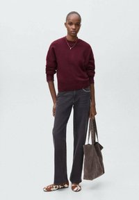 Eine Frau trägt einen burgunderfarbenen Pullover, dunkle Jeans, Schuhe mit Kuhfellmuster und hält eine große graue Wildleder-Tasche vor einem schlichten grauen Hintergrund.