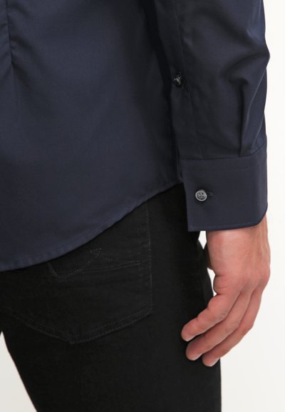 Chemise coloris bleu marine à manches longues, avec une texture lisse, un design ajusté, et un poignet boutonné, assortie à un jean noir avec une poche arrière.