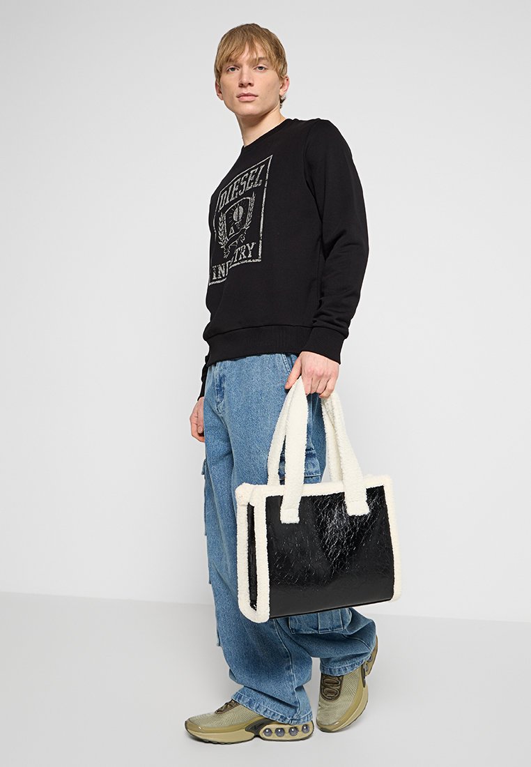 Giovane che indossa una felpa nera, jeans azzurri larghi e sneakers, mentre tiene una borsa tote nera con rifiniture bianche soffici, in piedi contro uno sfondo bianco.