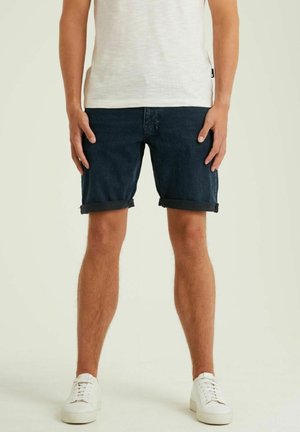 Un homme porte un short en denim bleu foncé, un t-shirt blanc et des baskets blanches, debout devant un fond clair uni.