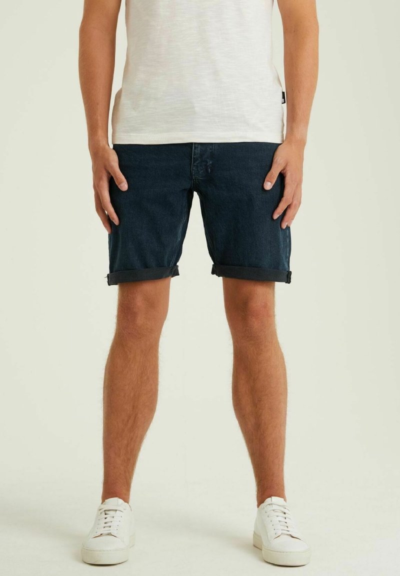 Un homme porte un short en denim bleu foncé, un t-shirt blanc et des baskets blanches, debout devant un fond clair uni.