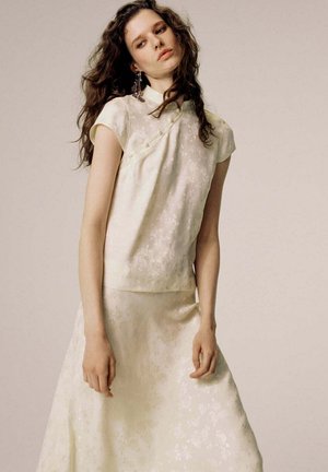 Mujer con pelo largo y ondulado, vestida con un top y falda texturizados de flores en crema, mirando ligeramente hacia abajo con expresión neutral.