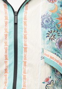 Valge jakk helerohelise tõmblukkude alaga, oranži „Just Cavalli” tekstiga tõmbluku kõrval ning värviliste lille- ja abstraktsete mustritega külgpaneelil.