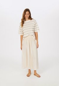 Gestreiftes Strickoberteil in Creme und metallischen Gold, kombiniert mit einem beigen Maxirock mit Smocking an der Taille und einem Seitenschlitz, ergänzt durch goldene Sandalen.