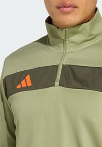 Šviesiai žalia sportinė maikutė su tamsiai žalia horizontalia juosta. Papuošta oranžiniais logotipo akcentais. Lygus paviršius, struktūrizuota apykaklė.