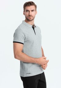 Šedé polo tričko s černým límečkem a manžetami, které má légu na dva knoflíky a vyšitým logem na lemu. Měkká textilie.
