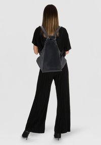 Donna con capelli lunghi e lisci, vestita con un completo nero e scarpe col tacco nere, che porta un grande zaino in pelle nera, voltata di spalle.