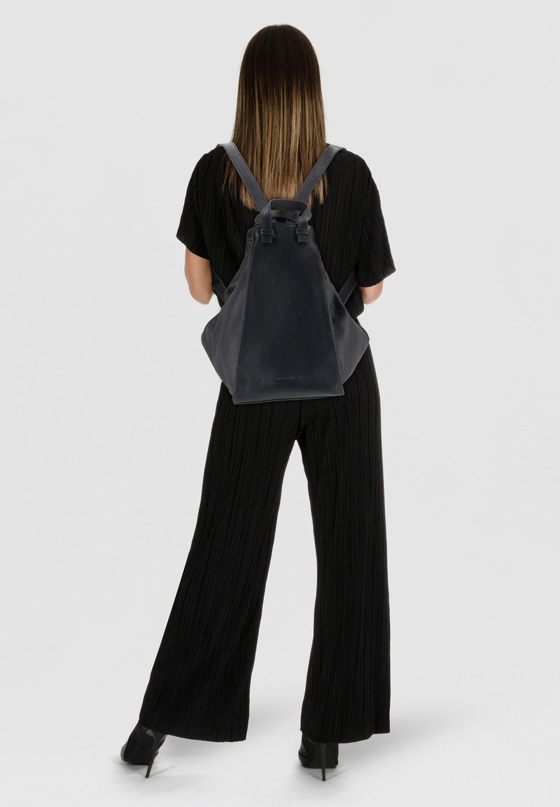 Donna con capelli lunghi e lisci, vestita con un completo nero e scarpe col tacco nere, che porta un grande zaino in pelle nera, voltata di spalle.