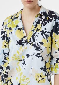 Blumenbluse mit hellblauem Hintergrund und gelben, schwarzen und sanft grünen Blumen. Sie hat einen Kragen und Dreiviertelärmel.