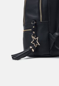 Schwarze, strukturierte Ledertasche mit goldenen Reißverschlüssen, Quasten-Details und einem sternförmigen Anhänger. Sie zeichnet sich durch ein definiertes Design und eine klare Form aus.