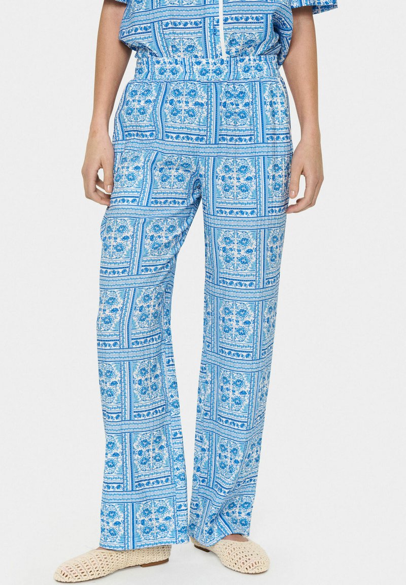 Šviesiai mėlynos spausdintos kelnės su gėlių ir geometriniu raštu, turinčios elastingą juosmenį ir plačias kojas, derinamos su nertomis béžinėmis bateliais.