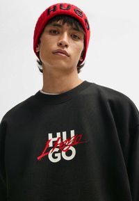 Felpa nera con logo "Hugo" bianco e rosso; berretto a maglia rosso con lettere nere. Texture liscia e vestibilità casual.