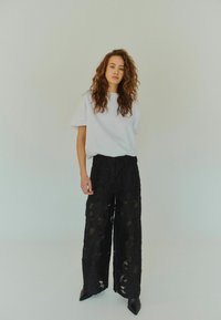 Bílý oversized tričko v kombinaci s černými krajkovými kalhotami s širokým nohavicemi, které mají průhledné výřezy. Model stojí na neutrálním pozadí a předvádí outfit.