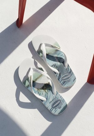 Paire de tongs blanches Havaianas avec un motif de vagues bleu et gris, posées sur une surface blanche sous une chaise rouge projetant des ombres.
